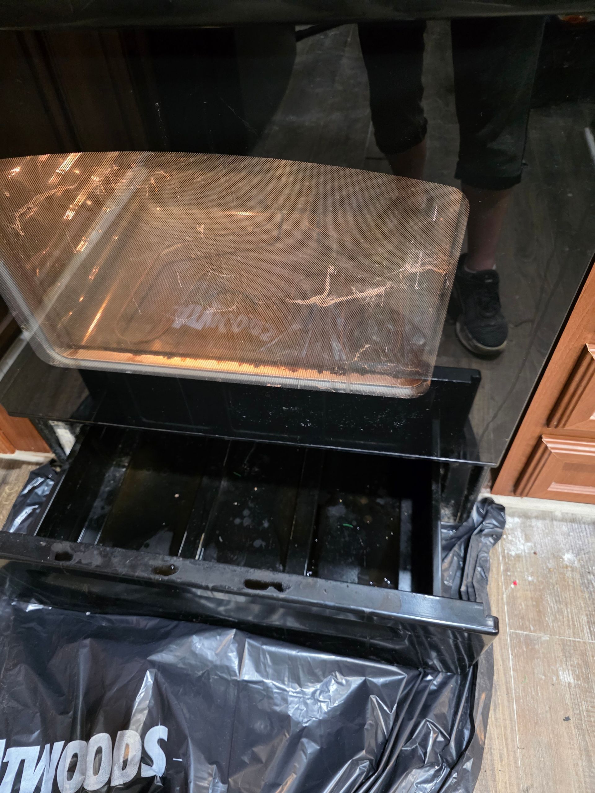 A person holds a dirty oven broiler pan over a garbage bag. Black surfaces and brown cabinets are visible.