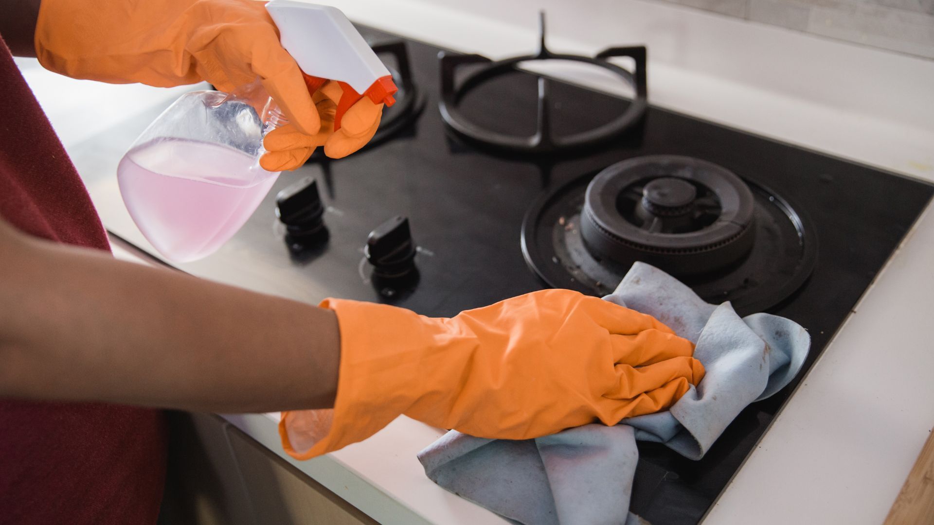 Hands in orange gloves cleaning a black stovetop with spray bottle and blue cloth.