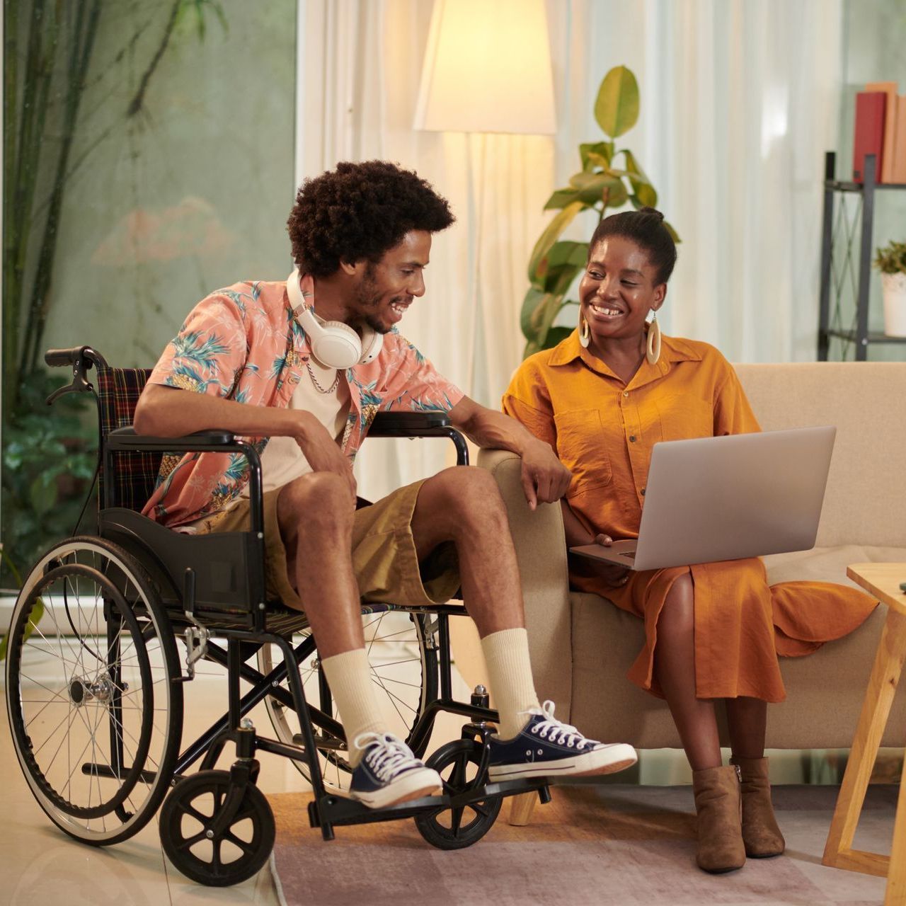 A man in a wheelchair is talking to a woman sitting on a couch with a laptop.