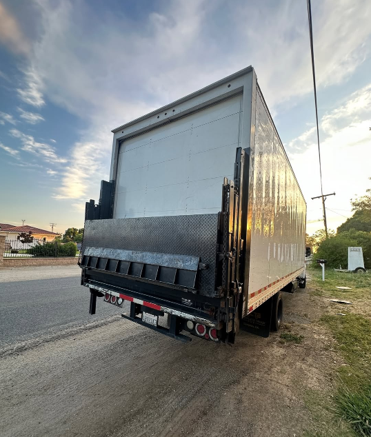 Rear view of a white box truck with a lift gate on a roadside with a cloudy sky in the background.