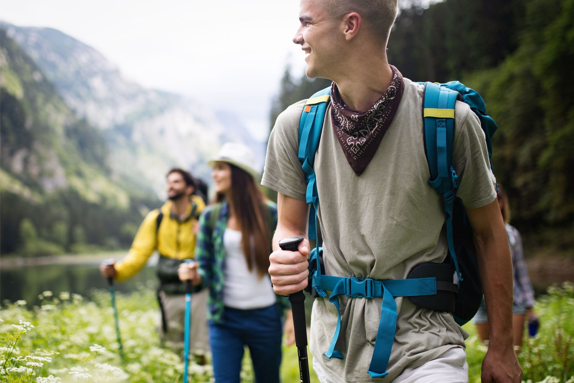 A person in the foreground hikes with a backpack and trekking pole, while two others walk behind them near a mountain lake.