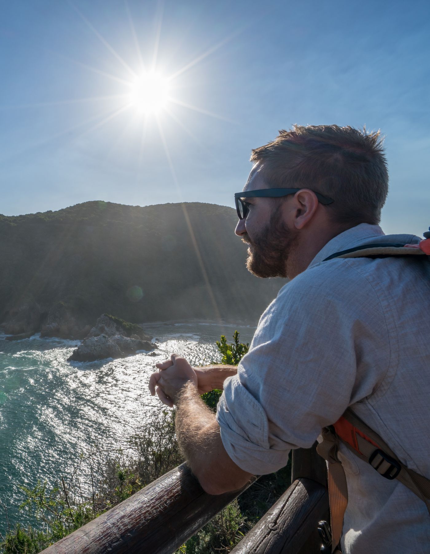 A hiker with a beard and sunglasses leans on a railing, looking out over a sunlit ocean and coastal hills.