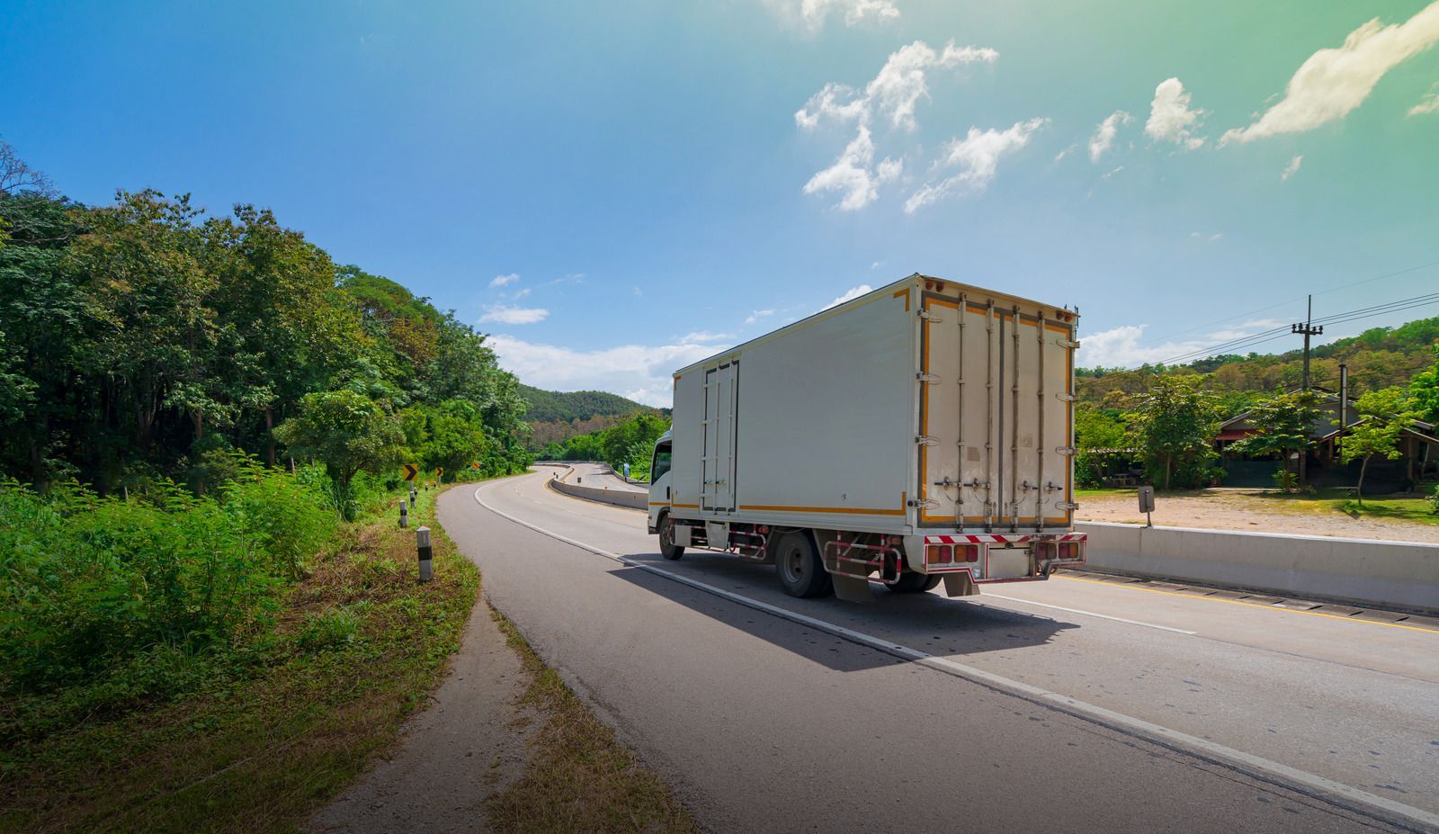 White cargo truck driving on a winding road through lush, green trees under a blue sky.