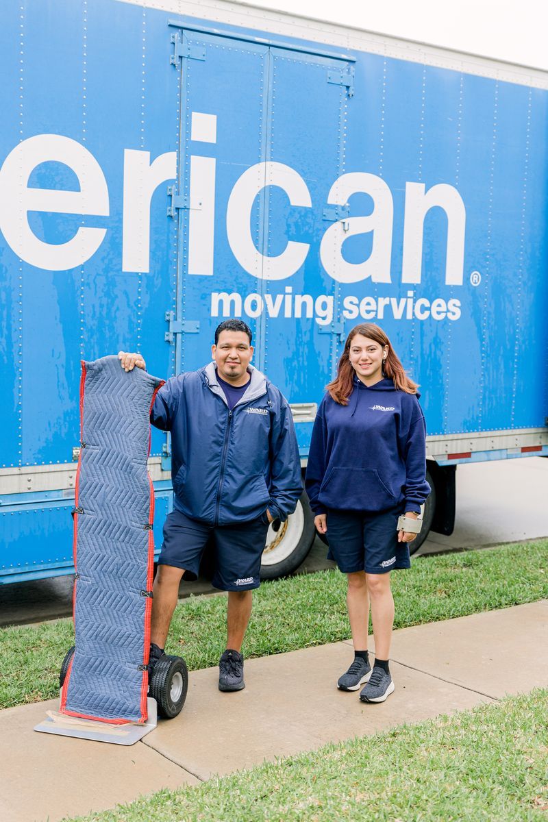Two people stand in front of an American Moving Services truck, one holding a hand truck.