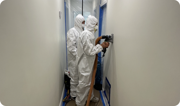 Two men in protective suits are working on a wall in a hallway.