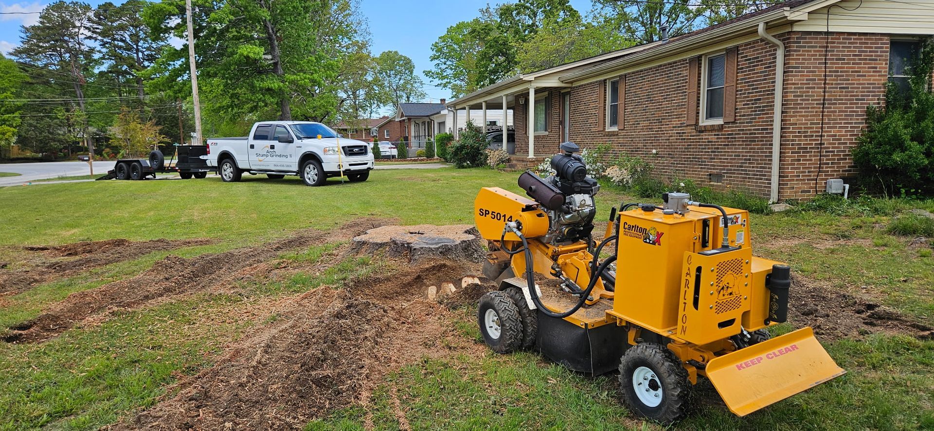 A yellow stump grinder is sitting in the grass in front of a house.