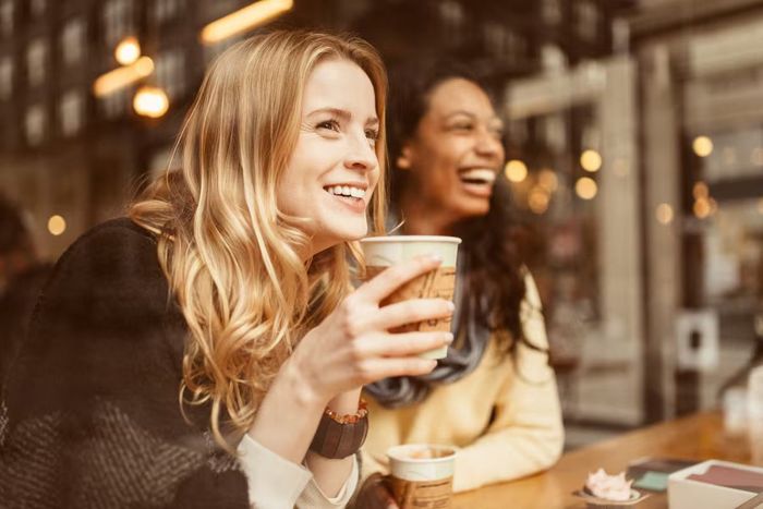 Two women are sitting at a table drinking coffee and laughing.