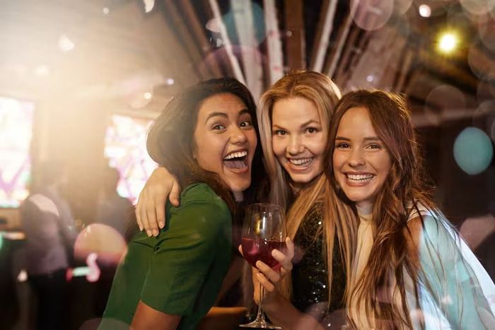 Three women are standing next to each other in a bar holding wine glasses.