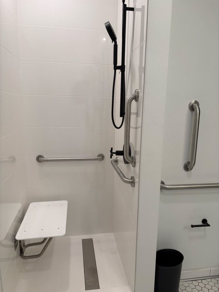 Accessible shower with grab bars, shower seat, and black showerhead in a white-tiled bathroom.