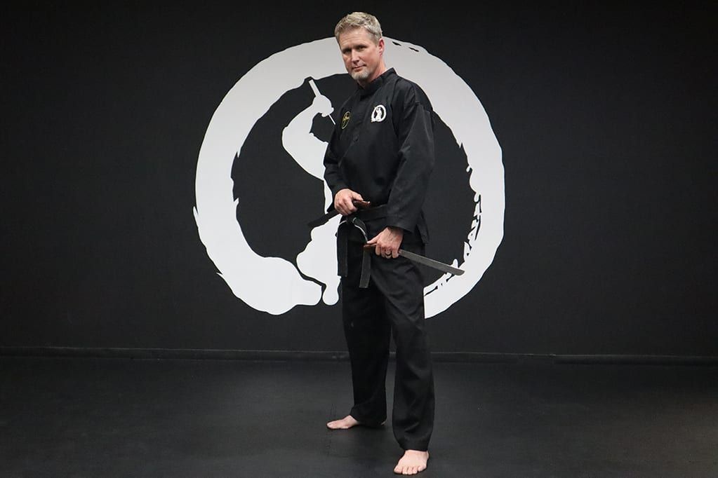 A martial arts instructor in a black uniform stands with nunchucks in front of a white emblem on a black wall.