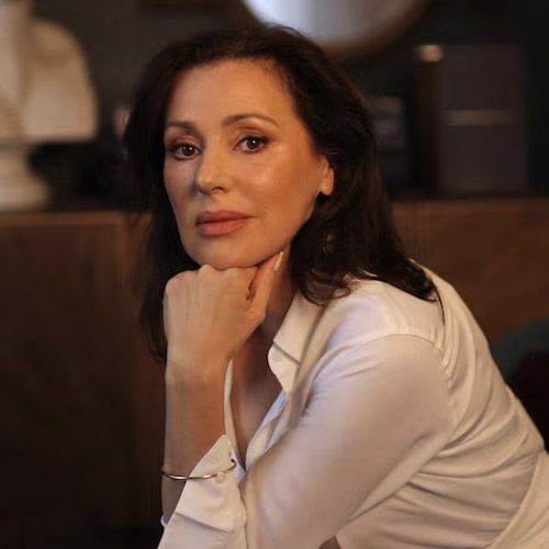 Tina Arena in a white shirt is sitting with her hand on her chin.