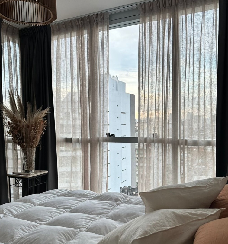 Dormitorio con cortinas transparentes de color beige, una gran ventana con vista a la ciudad