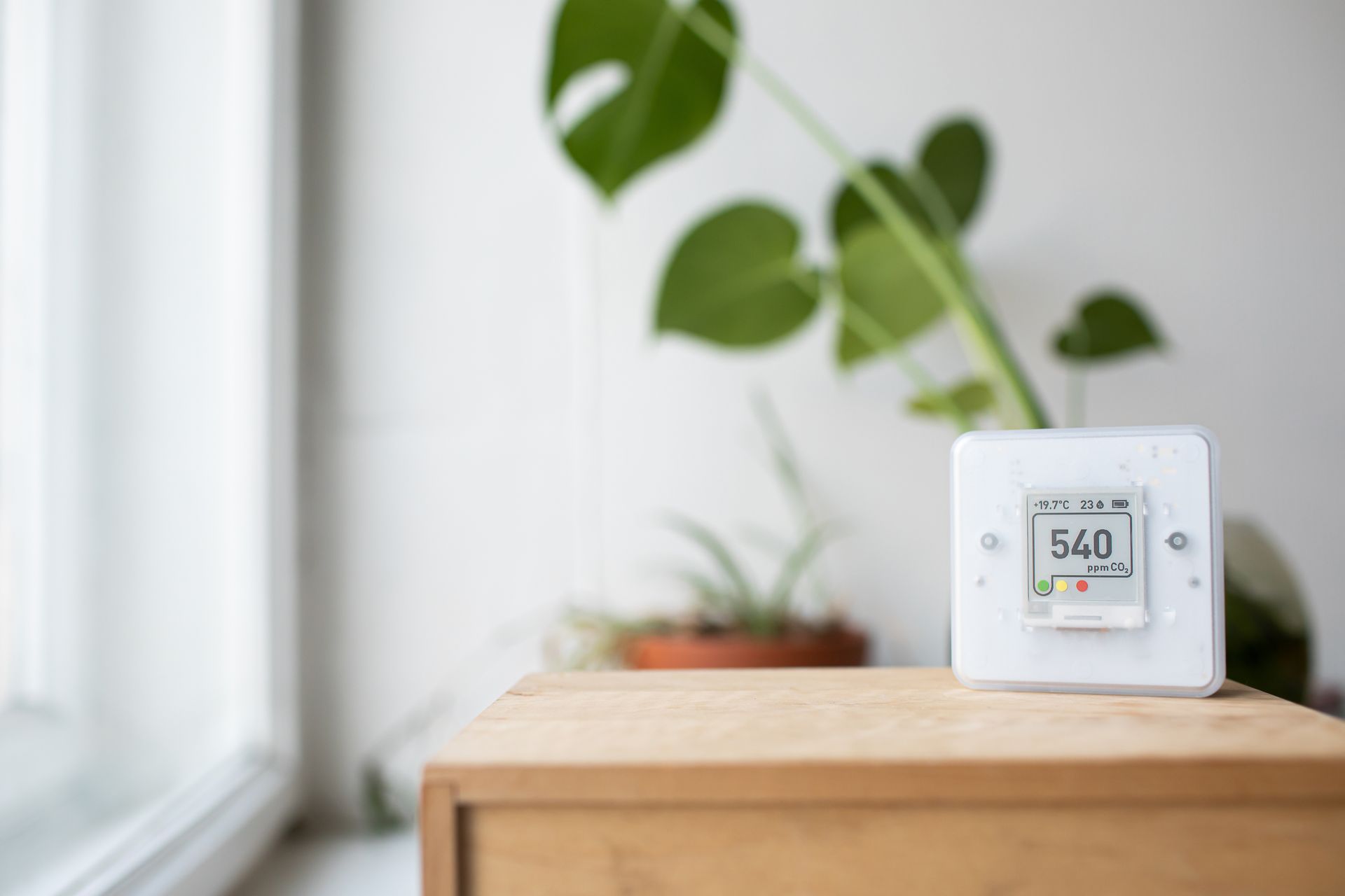 Un moniteur de qualité de l'air blanc affichant « 540 » est posé sur une surface en bois près d'une fenêtre et de plantes.