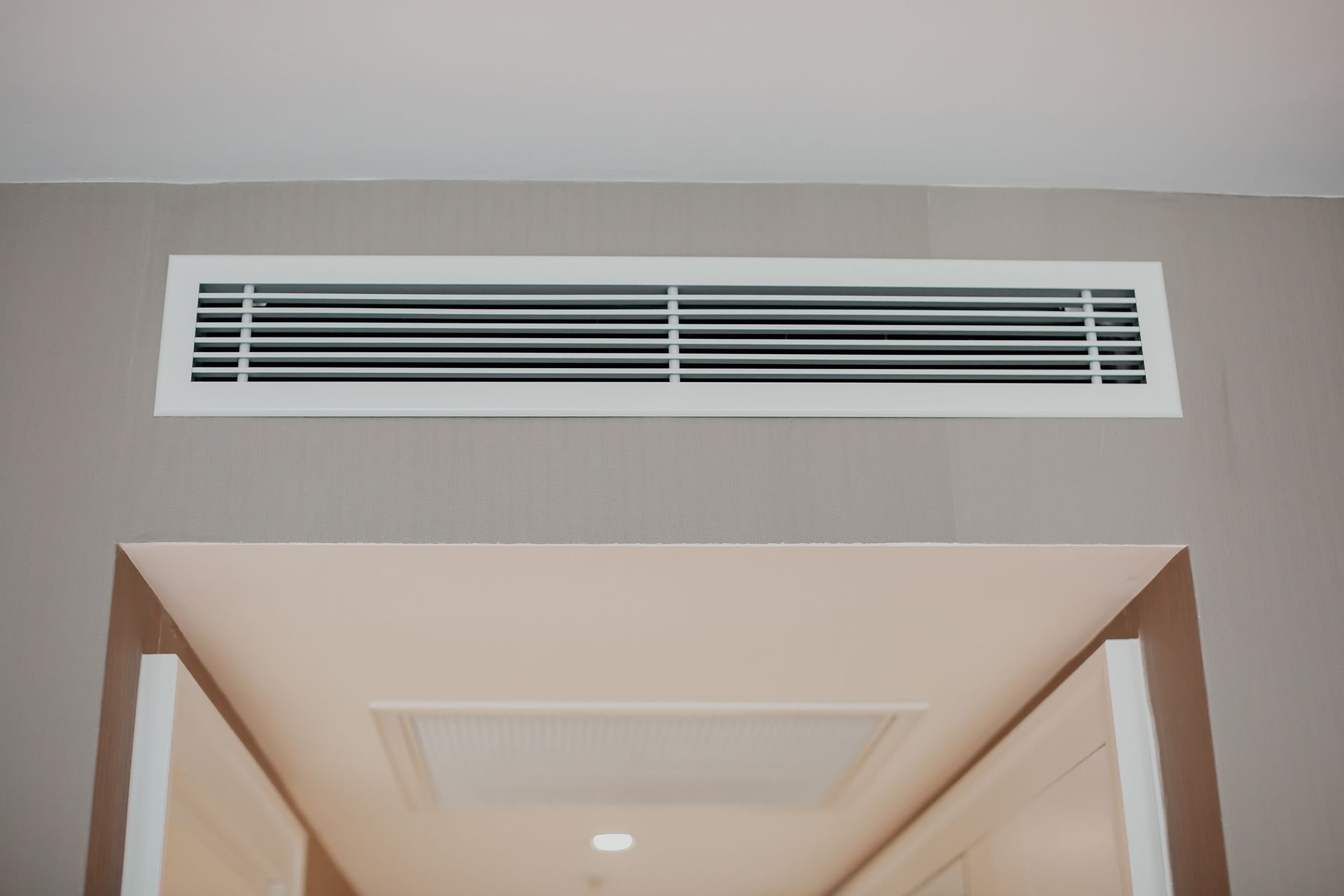 Grille d'aération blanche dans un cadre rectangulaire au-dessus d'une porte, encastrée dans un mur gris.