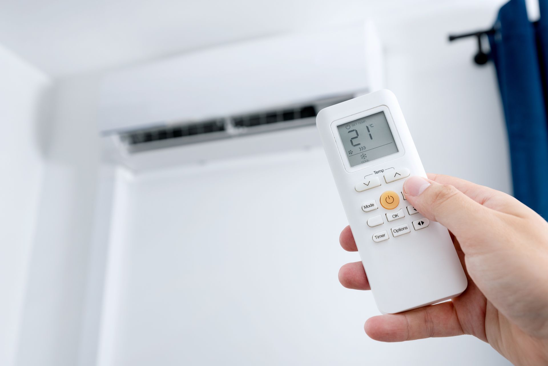 Main tenant une télécommande de climatisation, la pointant vers le climatiseur blanc sur le mur, réglé sur 21 degrés Celsius.