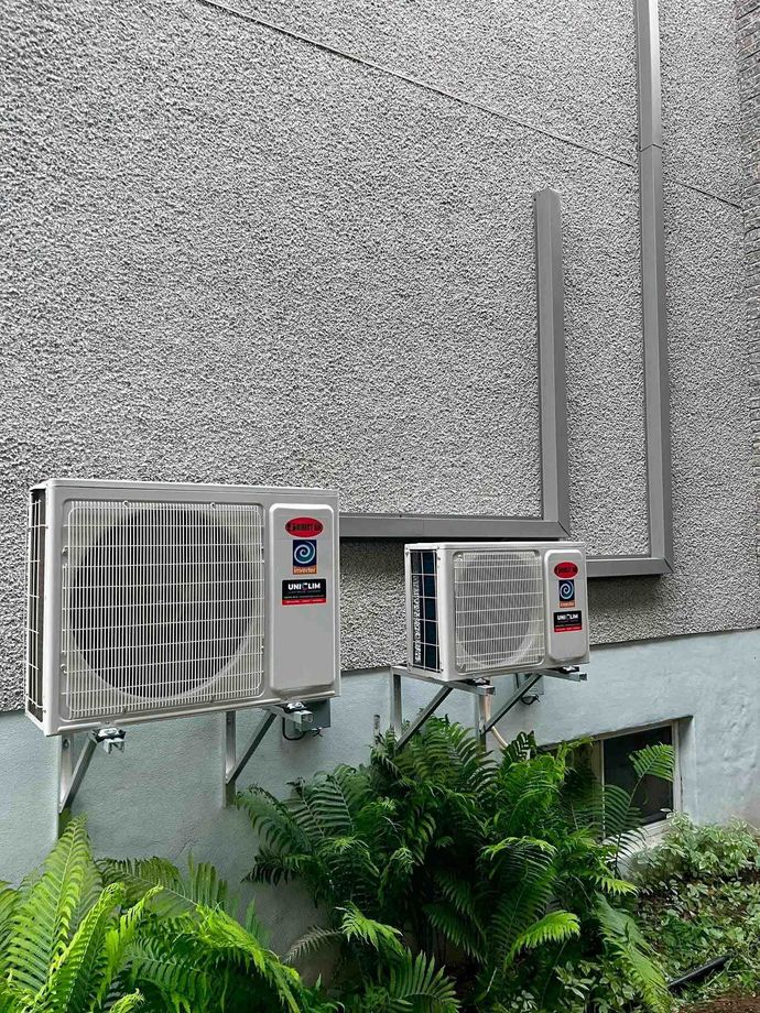 Trois climatiseurs extérieurs fixés sur un mur gris texturé. Des feuilles vertes sont visibles en dessous.