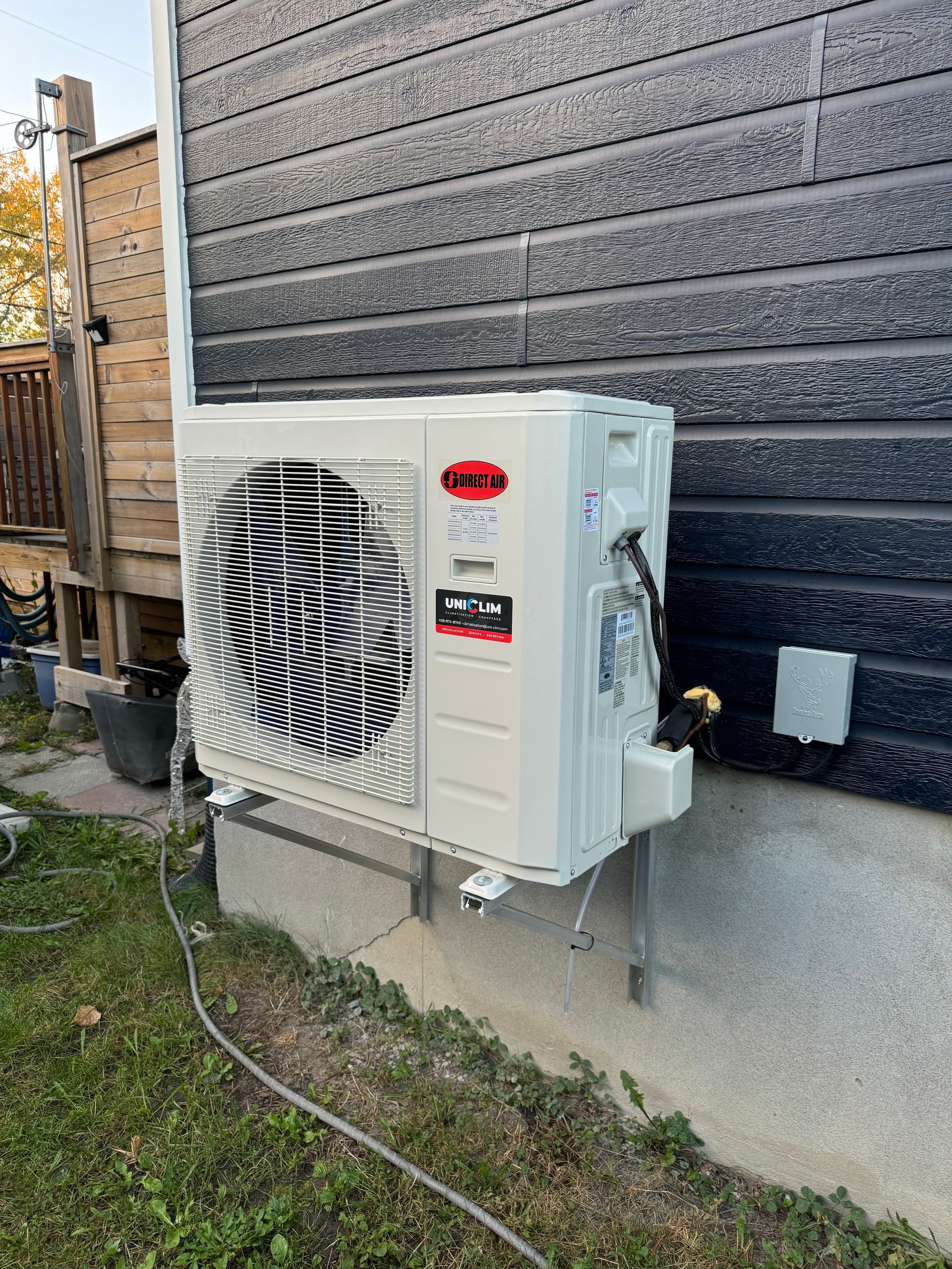 Unité de climatisation extérieure montée sur un bâtiment aux murs noirs, sur un terrain herbeux.