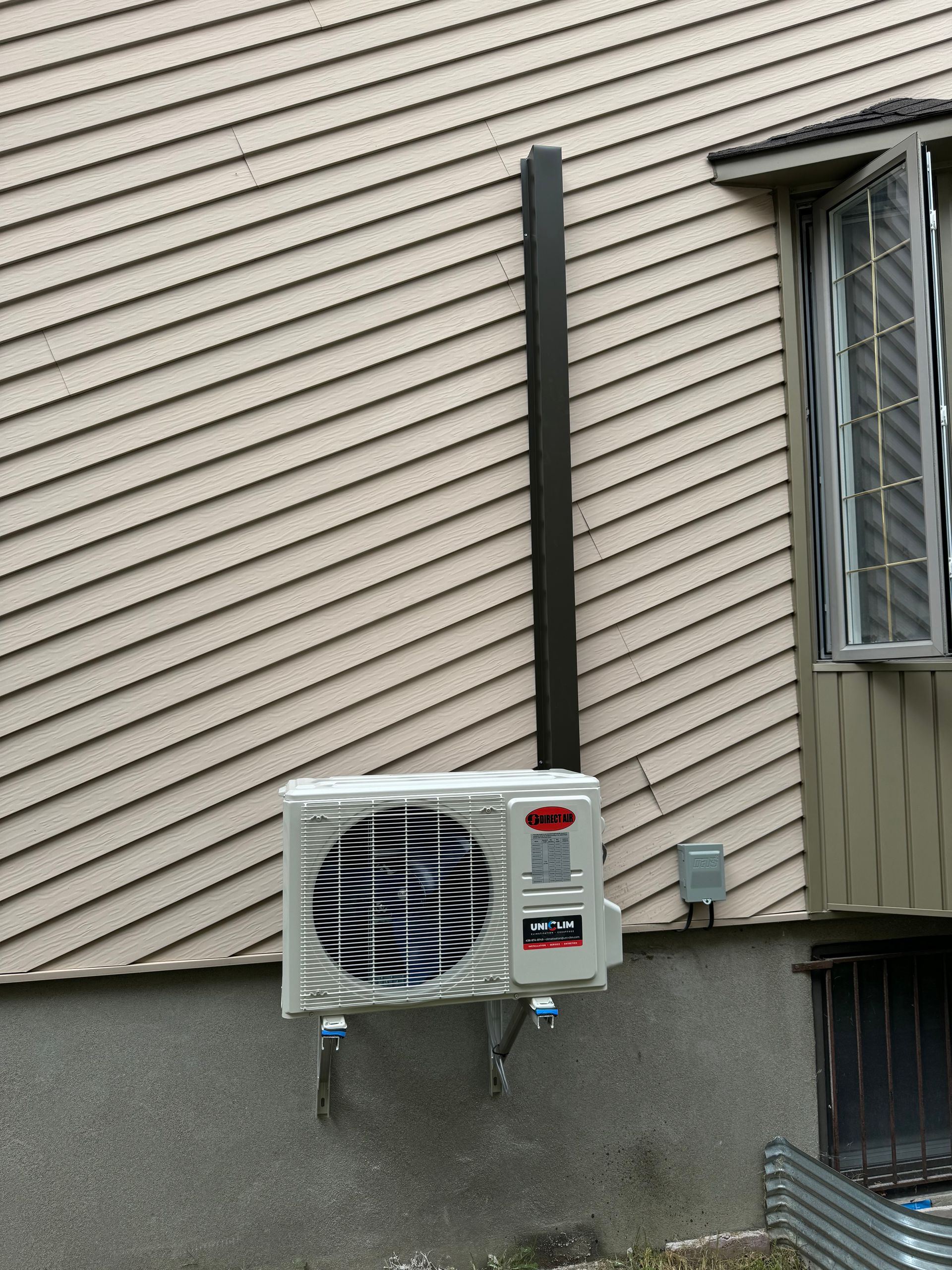 Un climatiseur extérieur est installé sur un mur beige. Un tuyau foncé court verticalement au-dessus de l'appareil.