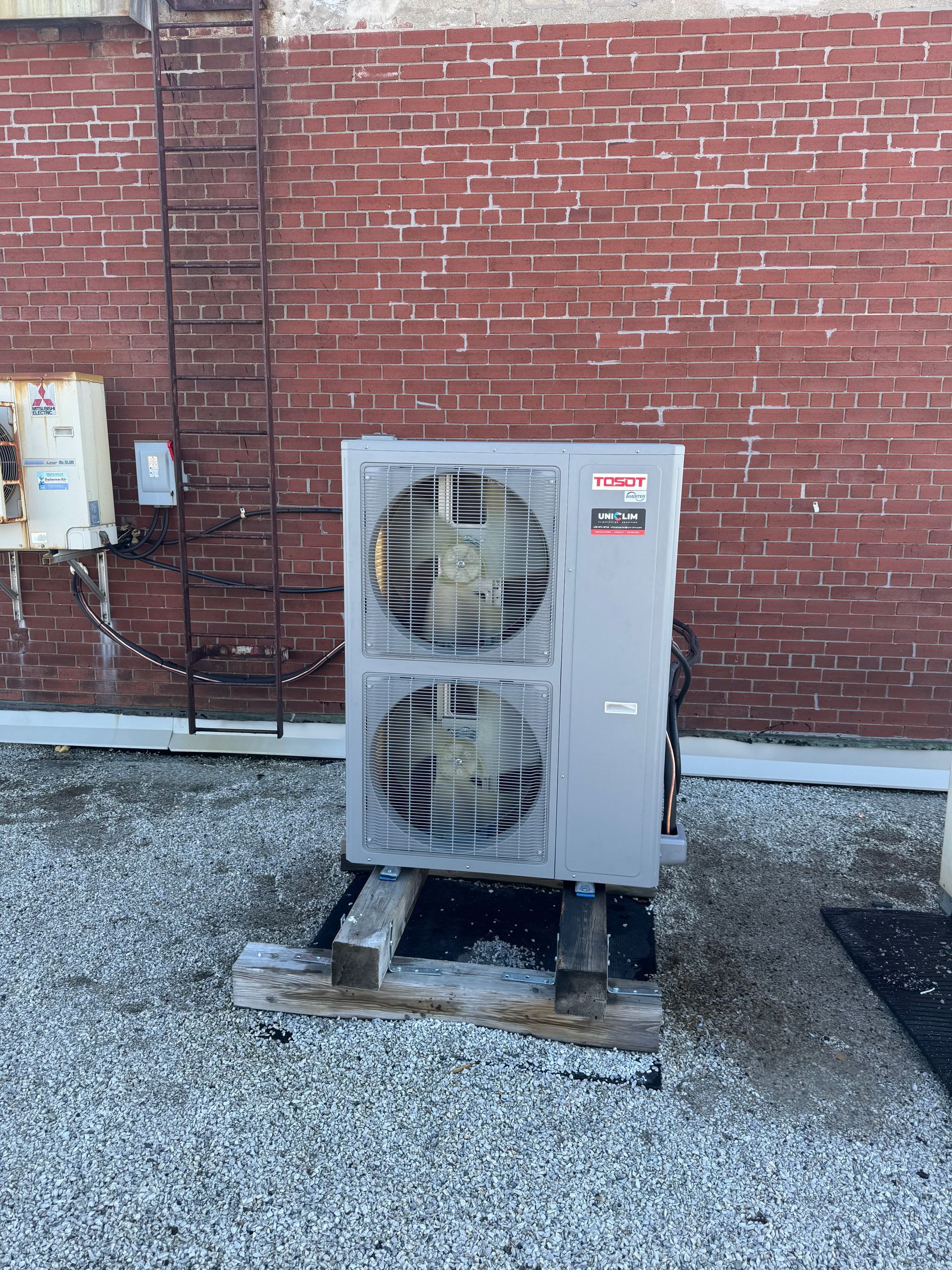 Unité de climatisation sur un socle en bois devant un mur de briques, avec une échelle métallique.