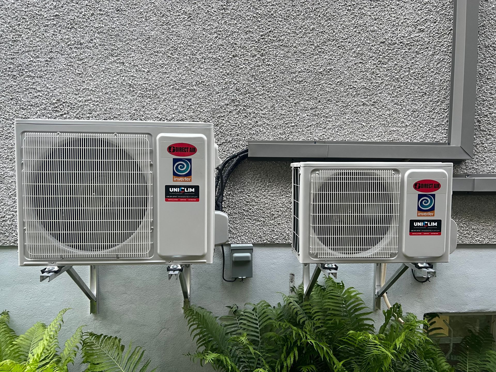 Deux climatiseurs extérieurs fixés sur un mur en stuc gris. Des plantes vertes luxuriantes se trouvent en contrebas.