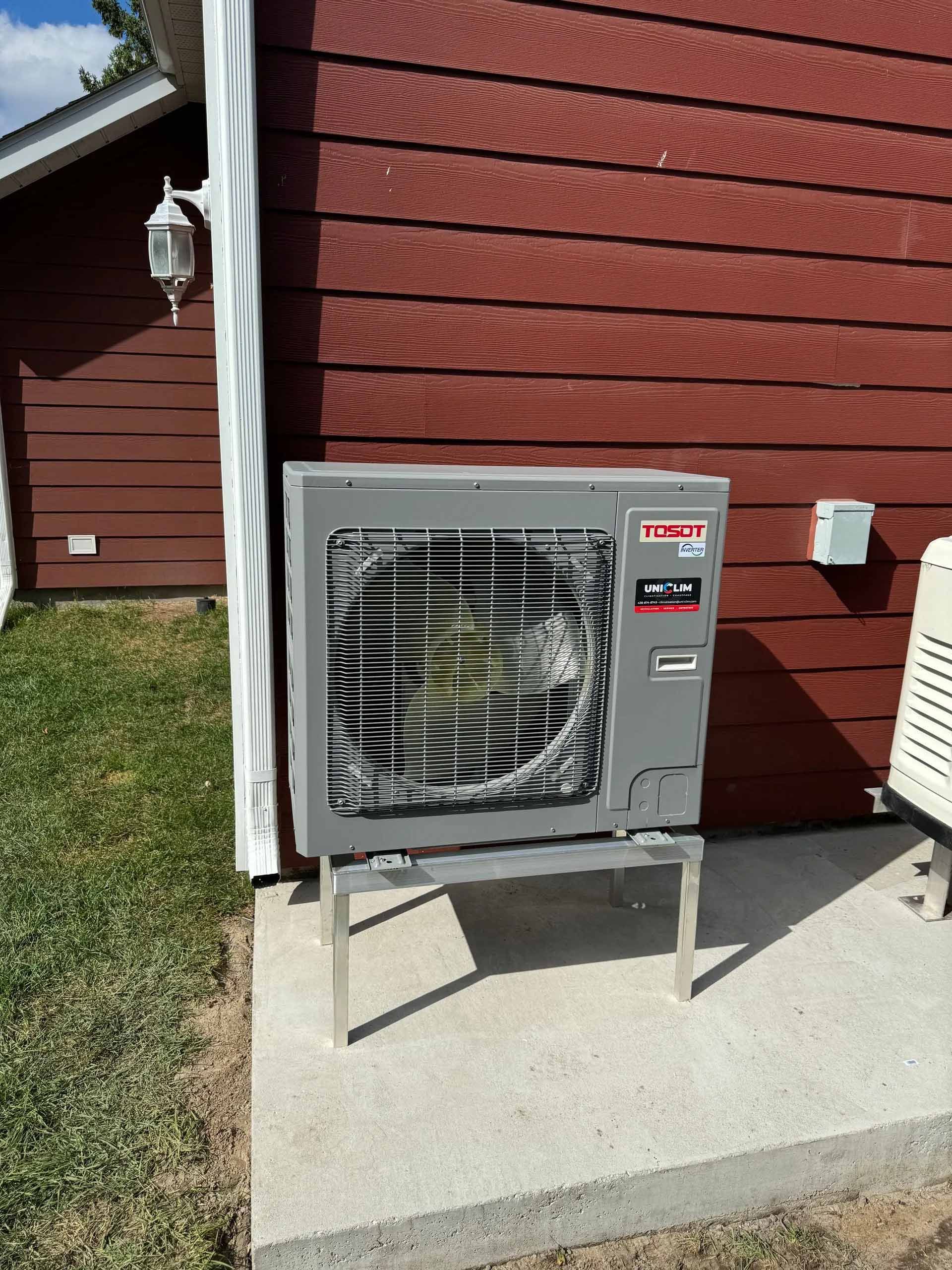 Unité de climatisation grise sur support argenté, à l'extérieur d'un bâtiment aux murs rouges, sur une dalle de béton.