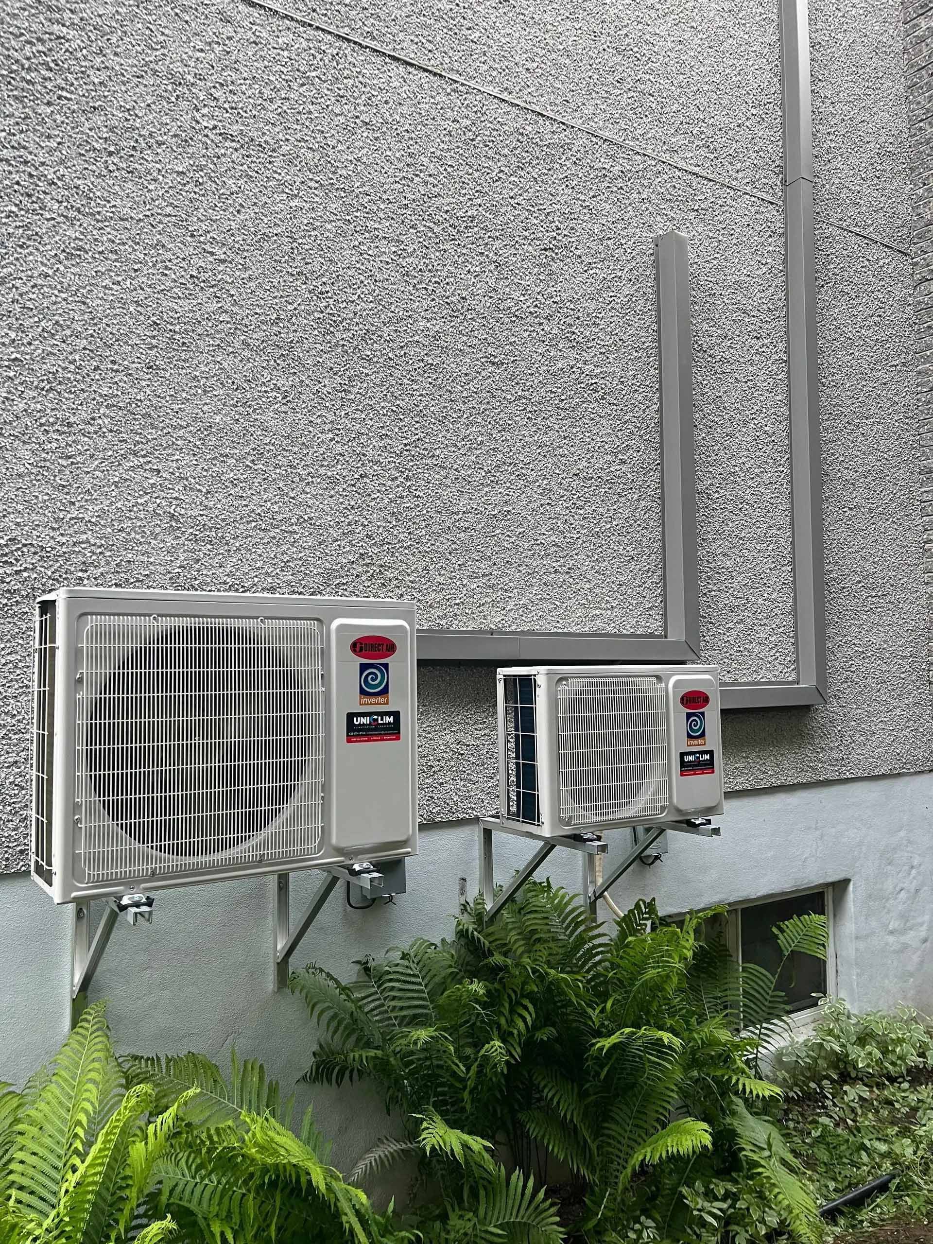 Trois climatiseurs extérieurs fixés sur un mur gris texturé. Des feuilles vertes sont visibles en dessous.