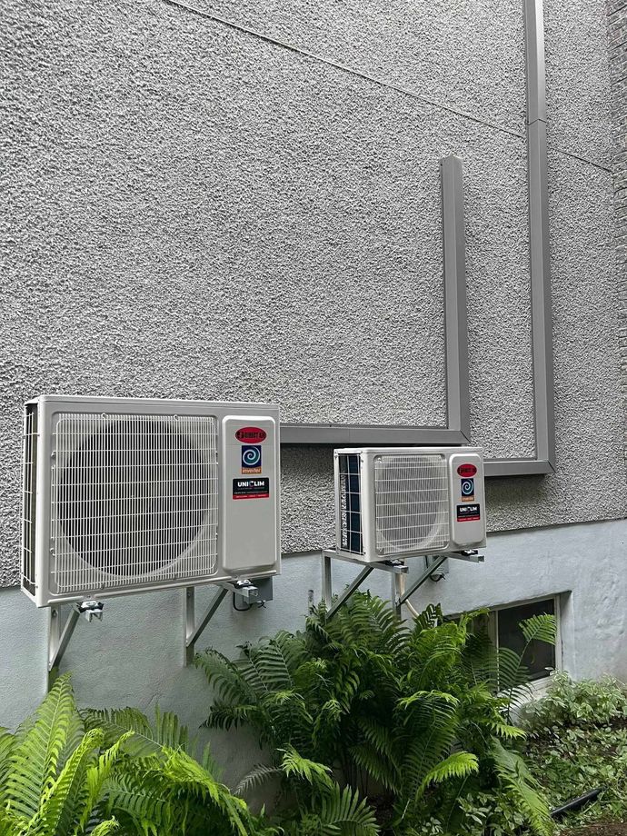 Trois climatiseurs extérieurs fixés sur un mur gris texturé. Des feuilles vertes sont visibles en dessous.