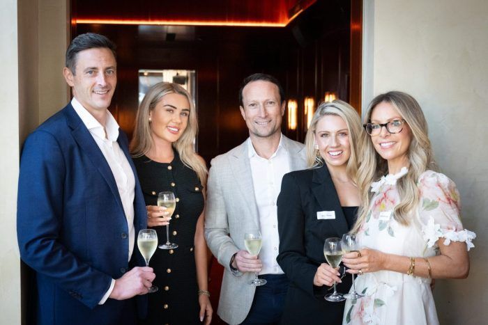 Five people dressed in business-casual attire stand in an indoor corridor, holding glasses of champagne and smiling.