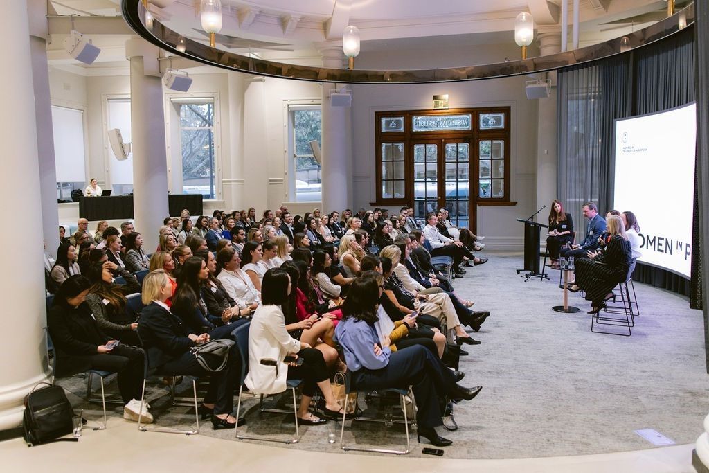 An audience sits in a bright, circular indoor hall watching a panel discussion on a stage with a large projection screen.