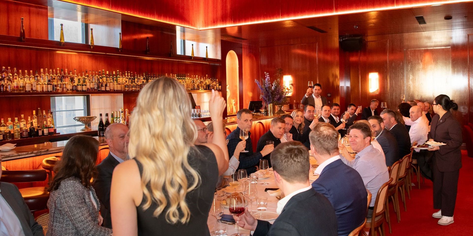 A group of people gathers for a formal dinner in a warm, wood-paneled restaurant with a large bar in the background.