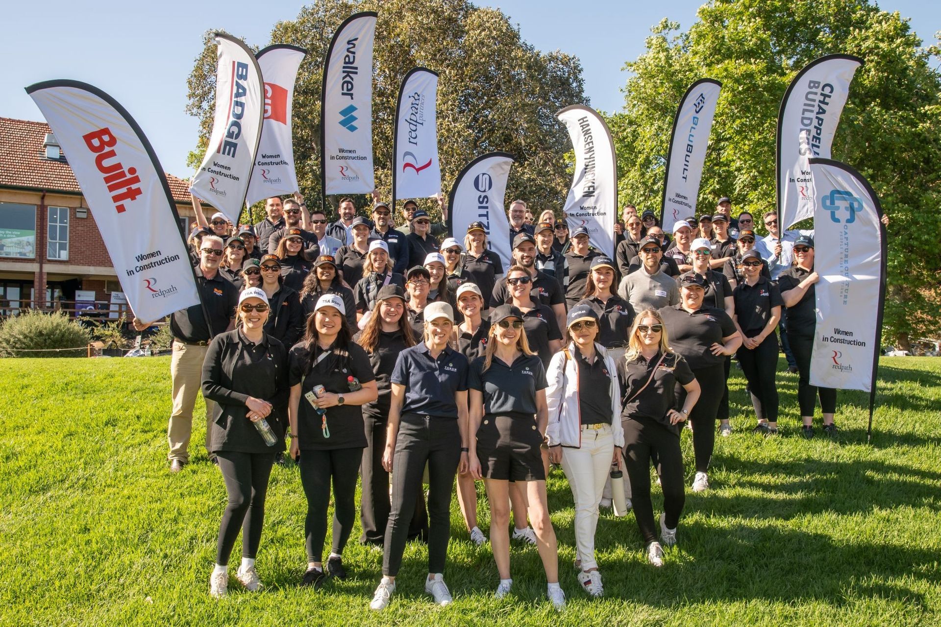 A large group standing on a grassy hill outdoors with sponsor banners, posing for a photo under a clear blue sky.