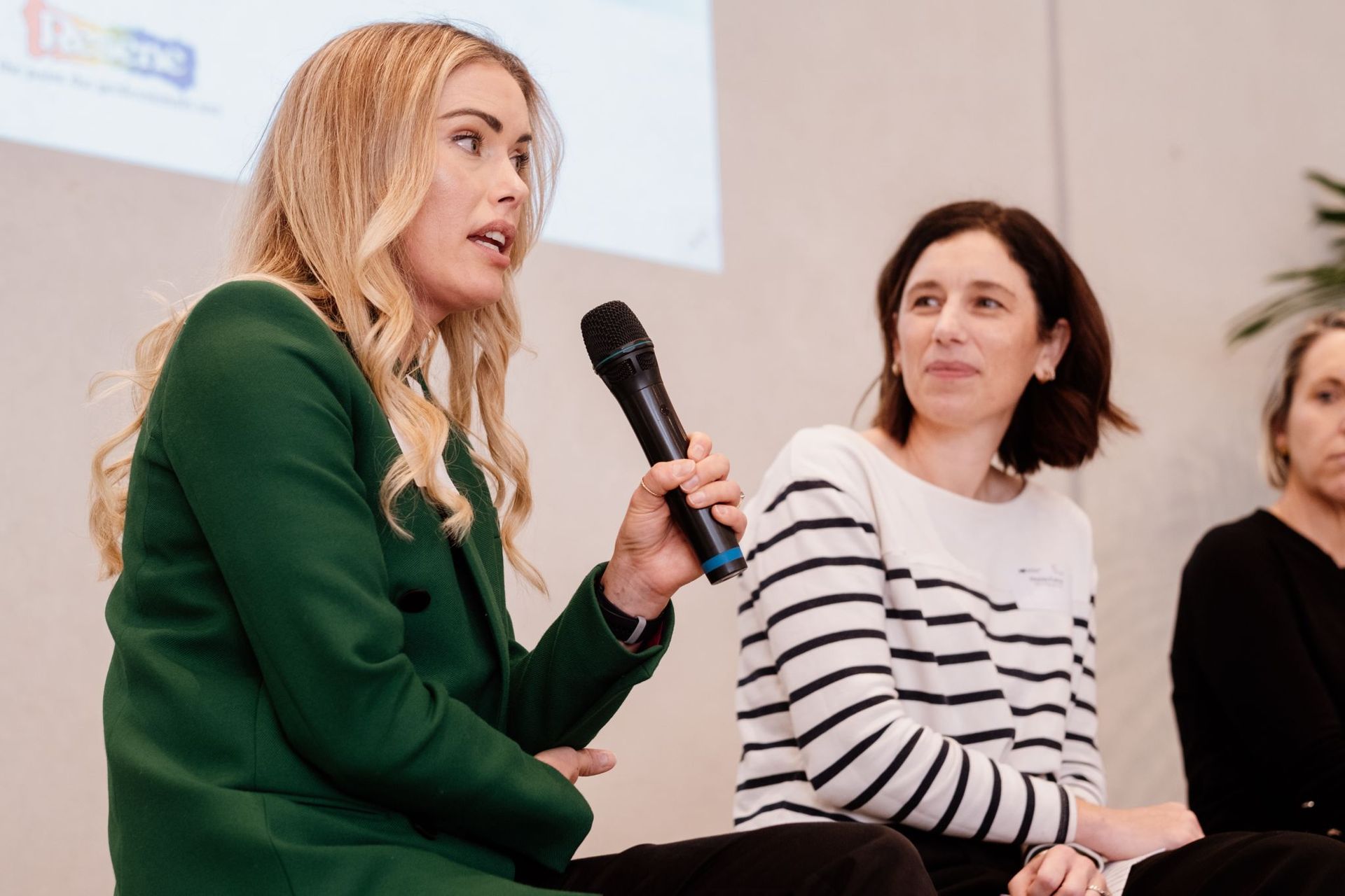 Three people participate in a panel discussion; one woman in a green jacket speaks into a microphone while others listen.