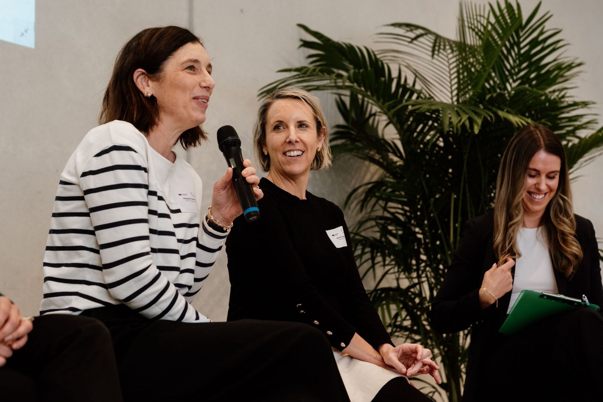 Three people sitting on a stage at an event; the person on the left speaks into a microphone, while others look on.
