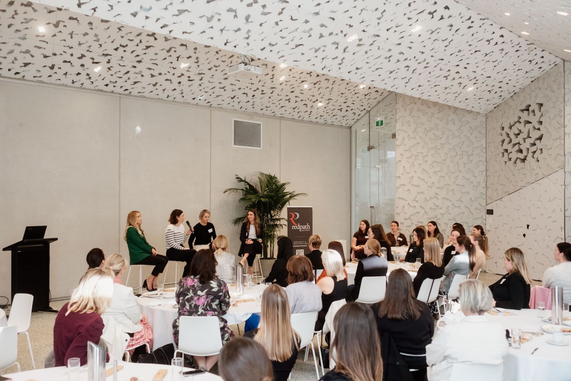A group of people listens to a panel of four speakers at a bright, modern event space with white circular tables.