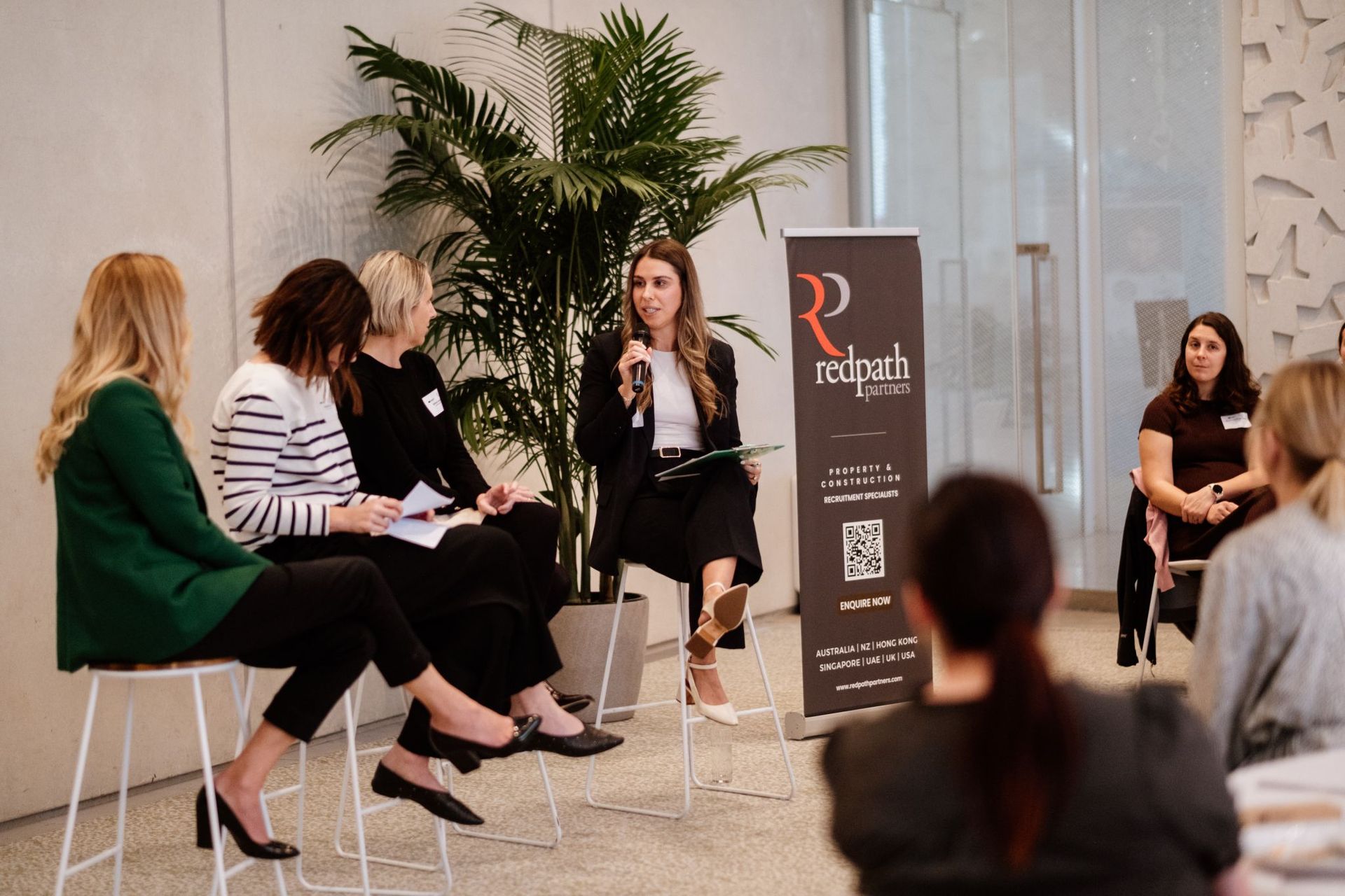 Four individuals on stools participate in a panel discussion near a large plant and a branded Redpath signage banner.