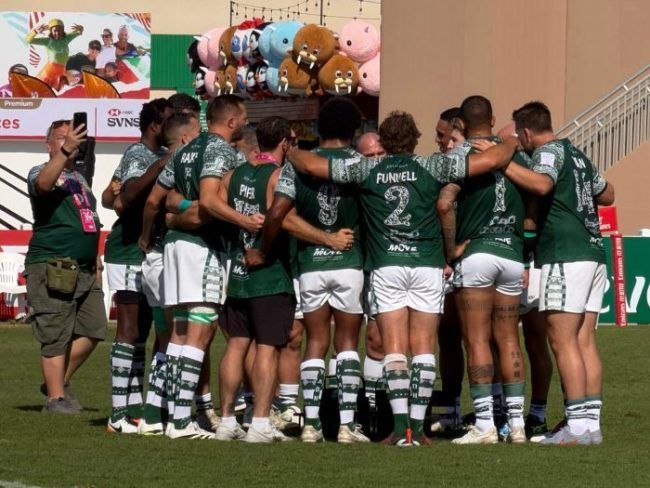 A group of rugby players in green and white uniforms huddled together on a field, with a spectator nearby filming them.