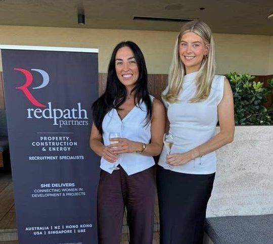Two women holding glasses stand smiling next to a Redpath Partners sign about women in property and construction.