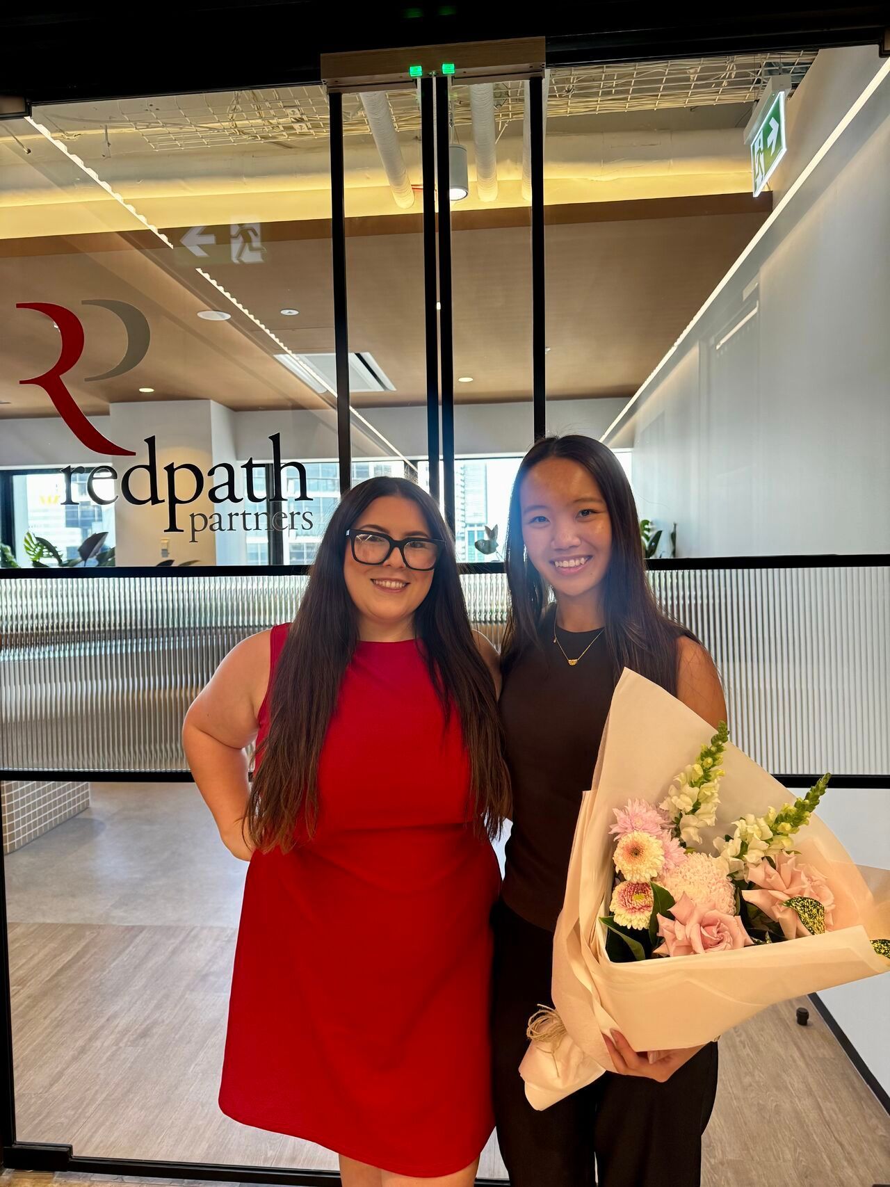 Two people smiling in front of a glass door with a