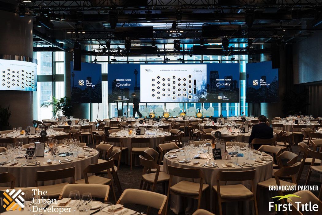 A banquet hall set for a business conference with round tables, chairs, and a large presentation screen.