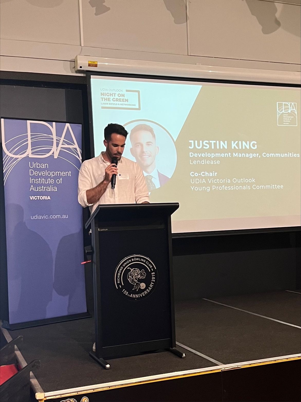 A speaker stands at a podium addressing an audience, with a projected slide showing his name, Justin King, and titles.