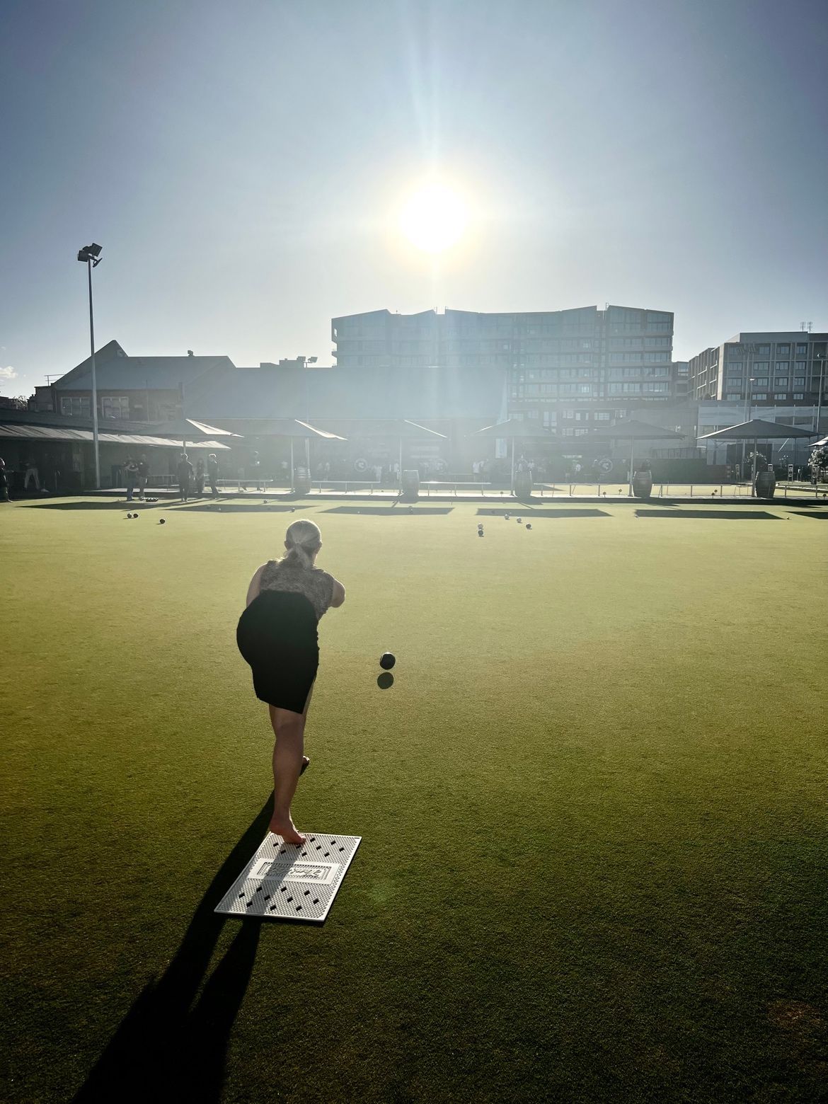 A person in silhouette bowls a ball on a green turf field at sunset, with buildings in the background.