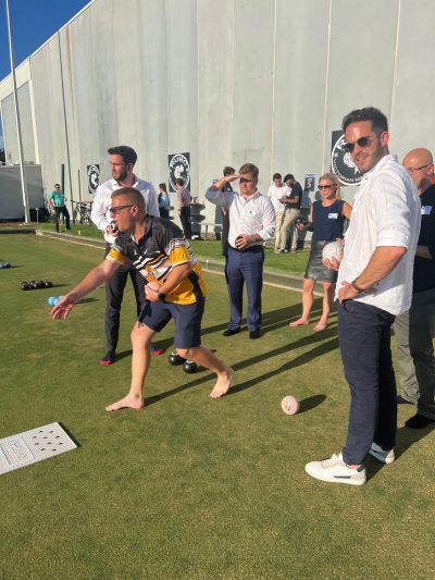 A group of people watch a barefoot individual bowling on a lawn green next to a large white wall.