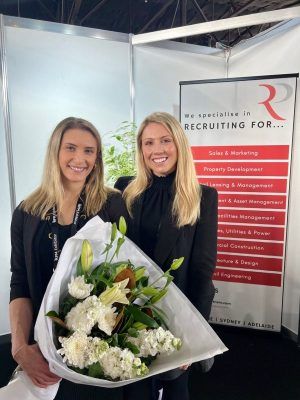 Two women stand together holding a bouquet of white flowers next to a vertical recruitment agency sign.