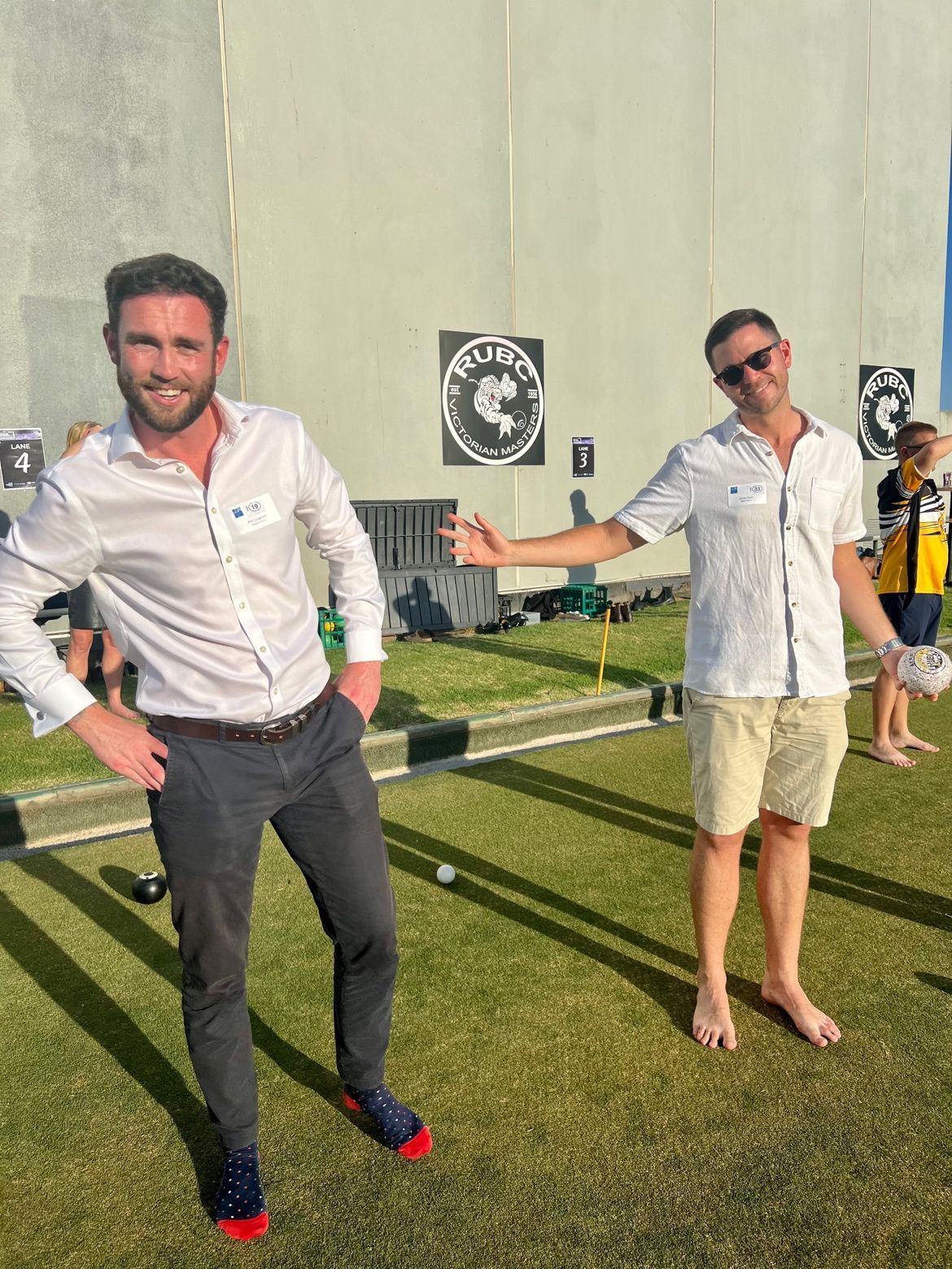 Two men in casual attire posing on a lawn near a wall with event signage, one standing and one gesturing toward a game.