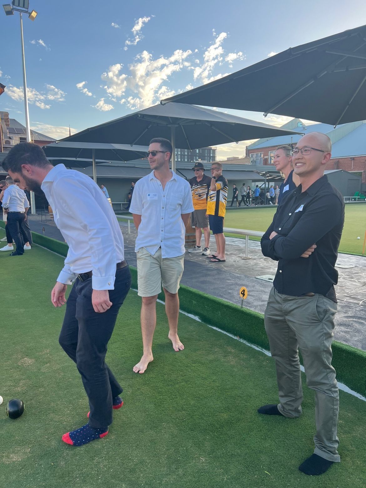 Three people play lawn bowls on an outdoor green under large umbrellas during the day.