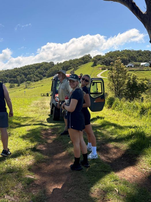 Two people stand in a grassy field next to an open utility vehicle on a sunny day with rolling green hills in the distance.