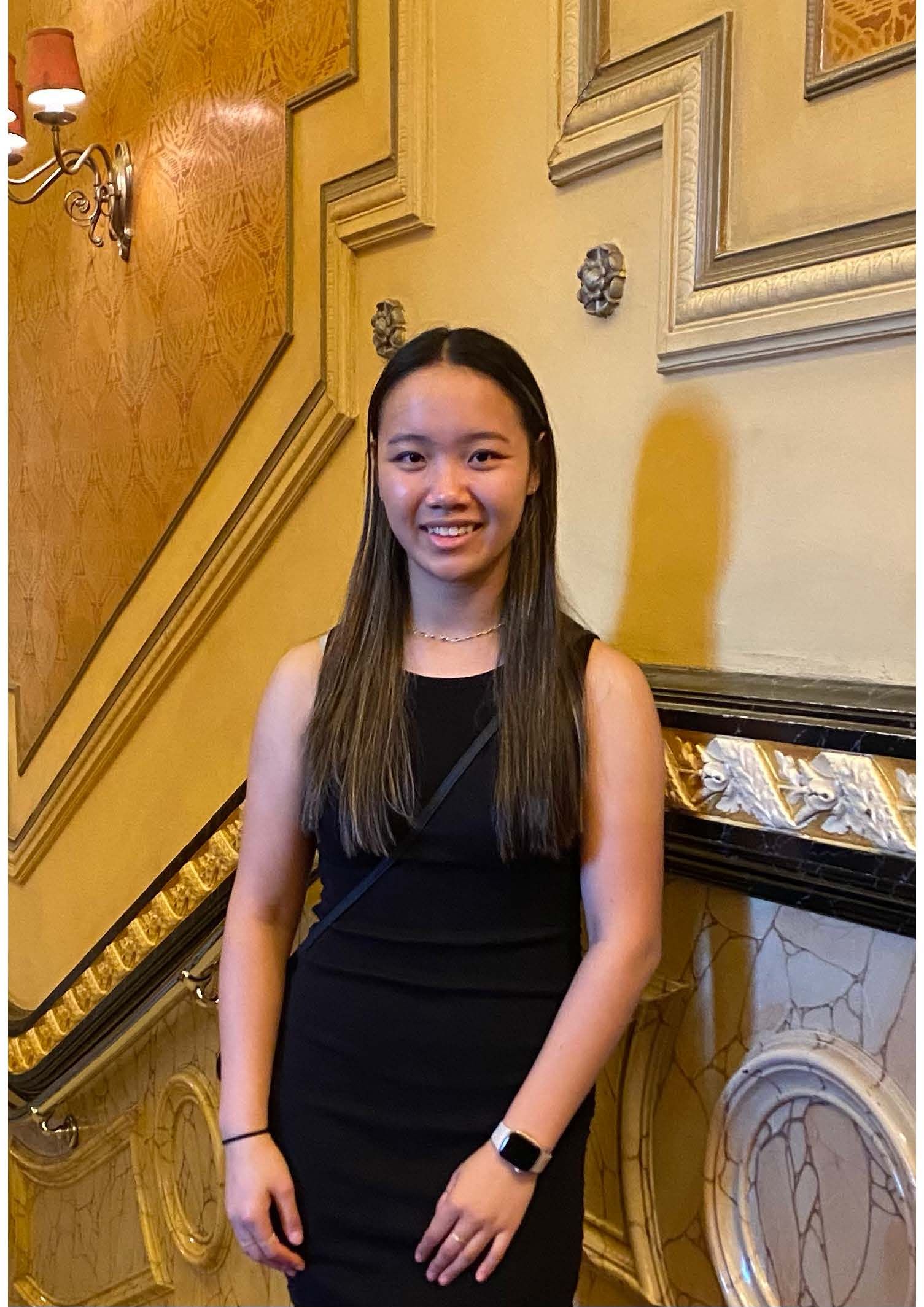 A smiling person wearing a black sleeveless dress and a watch stands in front of an ornate, golden-yellow wall.
