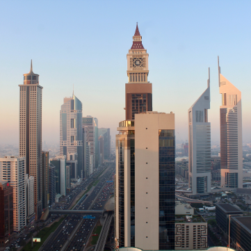 A sunrise view of Dubai's Sheikh Zayed Road, featuring tall skyscrapers, a clock tower, and traffic on the highway.
