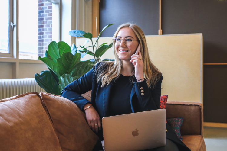 A smiling professional in a blazer talks on the phone while working on a laptop on a leather sofa in an office.
