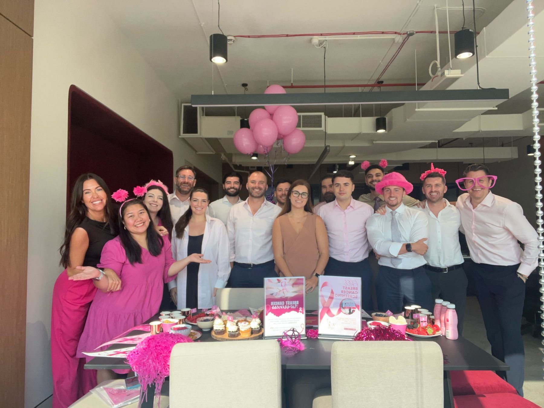 A group of colleagues wearing pink accessories gather in an office for a celebration with pink balloons and decorations.